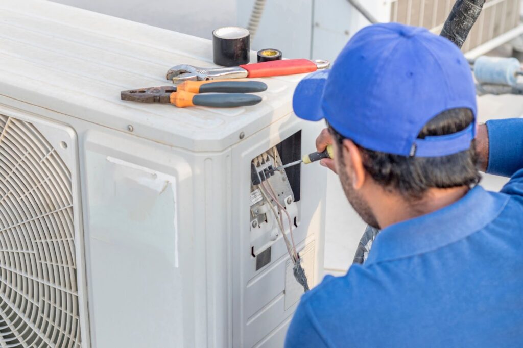 A technician in a blue uniform repairing an outdoor air conditioning unit using a screwdriver.