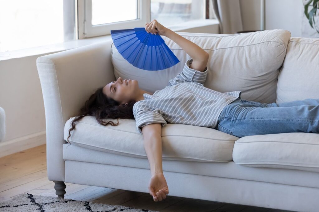 A woman lying relaxed on a light-colored couch in a bright room, holding up a blue folding fan to cool herself while her other arm hangs limply over the edge.